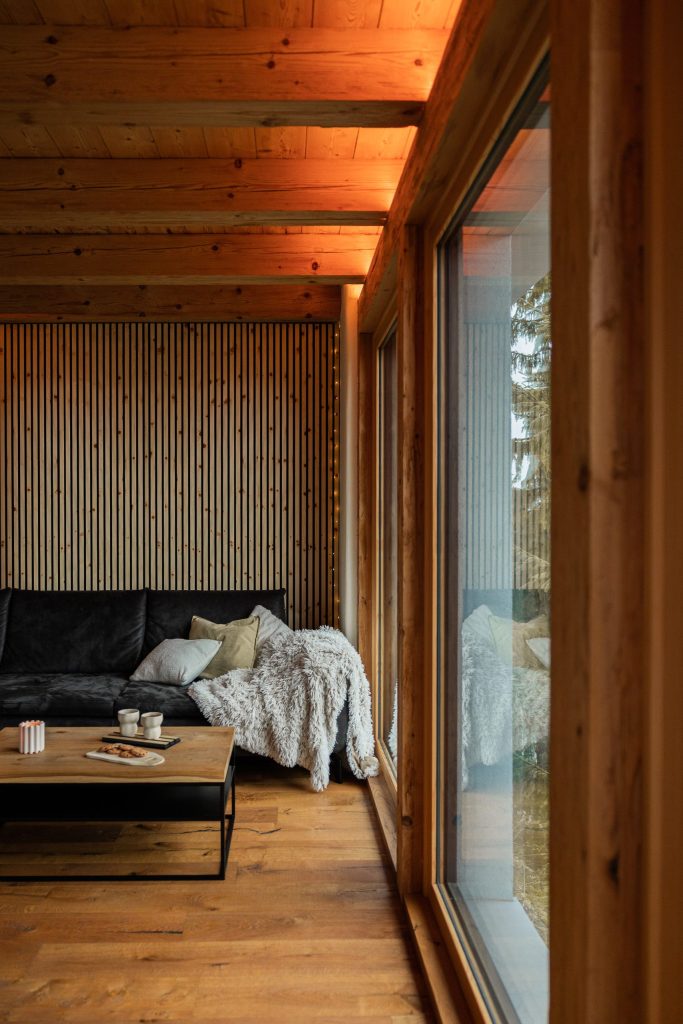 Gemütliches Wohnzimmer mit WoodUpp-Holzlamellenwand in vertikaler Optik, sichtbaren Holzdeckenbalken, warmem WoodUpp-Parkettboden und robusten WoodUpp-Holzfensterrahmen; schwarzes Samtsofa mit Kissen und flauschiger Decke, schlichter Holzcouchtisch mit Tassen und Gebäck, skandinavisch-minimalistischem Stil und ruhiger Atmosphäre.