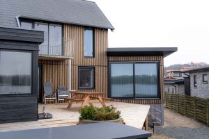 Moderne sommerhus med WoodUpp lodret facadebeklædning af smalle trælameller og WoodUpp terrasse med lyse træbrædder. På terrassen står et træ-picnicbord og to havestole; store mørke vinduesrammer, et lille metalgelænder og en plantekrukke i forgrunden og udsigt til vand i baggrunden.
