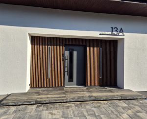 Indgangsparti med WoodUpp trælameller som varm, lodret facadebeklædning i naturlig træfarve omkring en mørkegrå dør, flankeret af to smalle indbyggede lyslister. Foran ses en lav WoodUpp indgangsplatform af store, grå, plankelignende fliser med mat, moderne finish, der kaster definerede skygger.