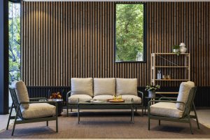 Stue med WoodUpp trælistevæg i mørkt fyrretræ og et WoodUpp-hyldesystem i lyst træ til højre. Foran ses en grønlakeret metalramme sofa og to lænestole med beige hynder, et lavt sofabord og dekorative glas på et brunt tæppe ved vindue med grønt udsyn.