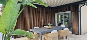 Udendørs terrasse med WoodUpps vertikale trælamelbeklædning i mørkbejdset træ og sorte alu-rammeprofiler omkring døråbning. Billedet viser også et gråt spisebord, fletstole med hvide hynder, en kurvekasse med grøn plante, tre kvadratiske sorte væglamper, glasskydedør og lyse fliser, store bananblade i forgrunden.