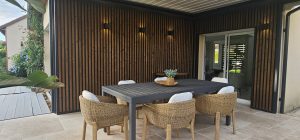 WoodUpp-lamelvæg med mørke, lodrette trælister i sort ramme pryder bagvæggen, flankeret af indbyggede væglygter. Billedet viser også WoodUpp-terrassebrædder til venstre, en grå udendørs spisebord, flettede stole med hvide hynder og plante centerpiece på flisebelagt terrasse, overdækket og med diskret LED-belysning.