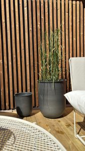 WoodUpp: Solbelyst terrasse med WoodUpps varme, lodrette trælameller med mørke mellemrum som bagvæg. To mørkegrå, matte WoodUpp urtepotter (en stor med højt prydgræs, en lille tom), delvist synlig hvid loungestol, rundt metalbord og lyst trægulv med vandpletter. Simpel, moderne æstetik.