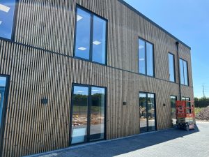 Moderne to-etagers erhvervsbygning med WoodUpp trælameller som vertikal facadebeklædning, mørke vinduesrammer med store reflekterende glaspartier, flisebelagt indkørsel og en rød sakselift parkeret ved siden af. Lamellerne er tynde, tæt stillede og skaber en lodret rytme på facaden; klar blå himmel og byggeplads i baggrunden.