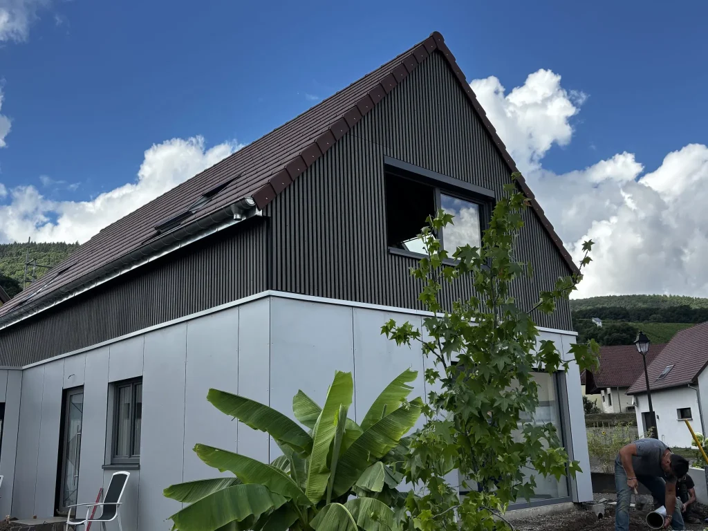Modernes Haus mit WoodUpp-Fassade: dunkel gerillte, schmale vertikale Holzlatten am Giebel und helle, hellgraue WoodUpp-Außenpaneele im Erdgeschoss. Offenes Fenster mit anthrazit Fensterrahmen, braunes Dach mit Regenrinne sichtbar. Bananenpflanze und junger Baum im Vordergrund; rechts ein Arbeiter, der Rohrarbeiten am Haus durchführt, unter blauem Himmel.