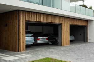 Moderne garagefacade beklædt med WoodUpp lodrette trælameller — smalle, varme brunlige facadepaneler med ensartede mellemrum omkring tre garageporte. WoodUpp-panelernes profil fremhæver et stramt, vertikalt design med synlige årer og mørke notspor, rammer portåbningerne ind og kontrasterer husets hvide overligger og glasaltan.