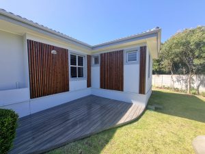 WoodUpp: Hjørne af hvidt hus med WoodUpp's lodrette, smalle trælamel-paneler i mørk bejdset finish monteret som facadebeklædning omkring vinduerne, samt WoodUpp gråt kompositdæk med let buet kant og tydelig træstruktur. Billedet viser også lille græsplæne, hæk og træer i baghaven.