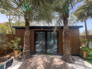 WoodUpp-haveskur beklædt med WoodUpp vertikale trælameller i varm brun finish, monteret med jævne mellemrum som slat-facade og afskærmning. Sort aluminiumskarm om store glas-skyderdøre og mørkt tagudhæng. To palmer står i forgrunden, pottede planter, lyskæde over indgangen og træhegn langs siderne, med pool i baggrunden.