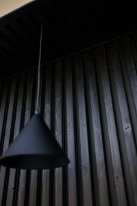 Mørkt, minimalistisk foto af WoodUpp: lodrette, sorte/brune shou-sugi-ban lignende træpaneler med synlige åretegninger, knaster og skruer, dybe riller og mat kulsort overflade. Forgrundens matte, koniske WoodUpp-pendel hænger i tynd ledning under et mørkt udhæng, skaber et moderne, sobert udtryk med bløde skygger og kontrast.