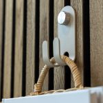Foto zeigt WoodUpp Holzlamellen-Wandverkleidung in Eichenoptik und einen hellgrauen WoodUpp Metall-Haken mit geriffelter Schraube, an dem ein Korbgriff und ein Buch („WHITE & FADED“) hängen. Detaillierte Holzstruktur, schlanke Hakenform und mattierte Metalloberfläche sind klar sichtbar. Nahaufnahme betont die natürliche Maserung und filigrane Verarbeitung.