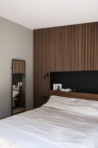 Schlafzimmer mit WoodUpp-Holzlamellenwand und WoodUpp-Kopfteil: gleichmäßig angeordnete, dunkelholzfarbene vertikale Lamellen, eingebautes schwarzes Kopfteil-Regal mit Ablageflächen, schwarze Leselampe und schmaler, rahmenloser Ganzkörperspiegel. Auf dem Regal liegen Bücher und eine skulpturartige Dekoration; weiß bezogenes Bett im Vordergrund, mit sichtbarer Holzmaserung und ruhiger Farbpalette.