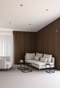 Minimalistisches Wohnzimmer mit WoodUpp-Holzlamellen: dunkle, schmale, vertikal montierte Wandverkleidung von WoodUpp mit feiner Struktur und klaren Linien, auf zwei Wänden in der Zimmerecke installiert, die die Architektur betont. Dazu helle beige Sofas, schwarze Kubus-Beistelltische, weiße Lamellenjalousien und heller Teppichboden.