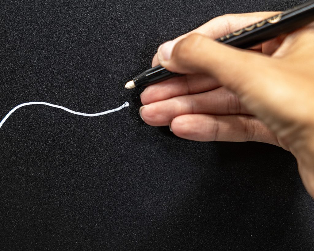 WoodUpp: Eine Hand hält einen schwarzen Kreidemarker von WoodUpp mit heller Rundspitze und zeichnet eine weiße, geschwungene Linie auf eine matte schwarze Tafel. Das glänzende Stiftgehäuse, die kräftige weiße Tinte und präzise Linienführung sind im Foto gut erkennbar.