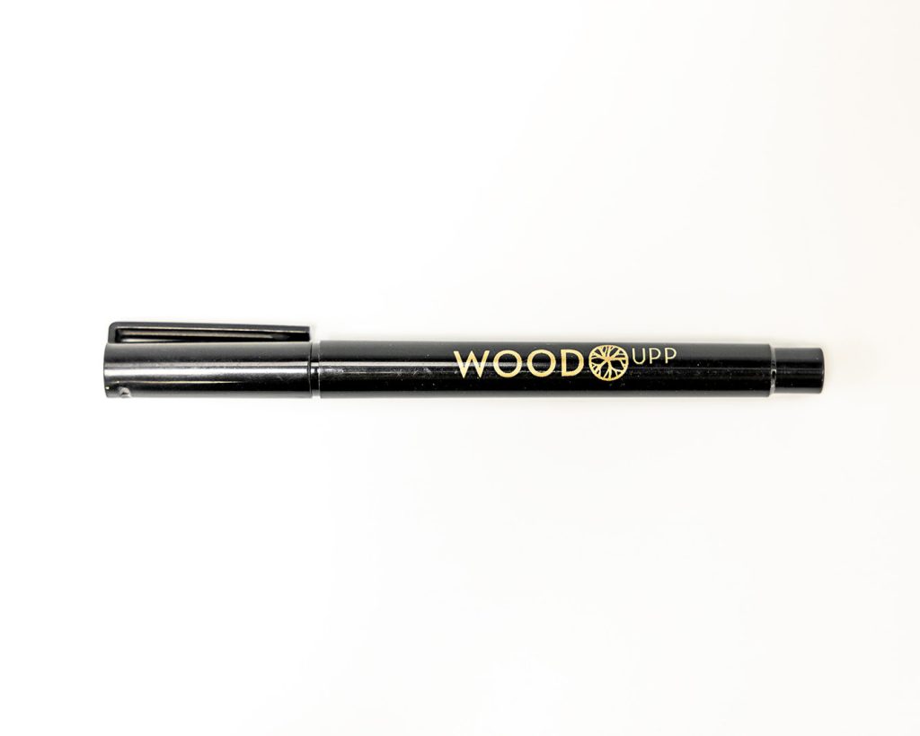 Schwarzer WoodUpp Filzstift mit abnehmbarem Clip-Deckel liegt horizontal auf weißem Hintergrund. Der Deckel sitzt links, das Stiftende rechts. Auf dem Stift prangt das goldene WoodUpp-Logo mit Rad-Emblem in glänzender Metallic-Prägung. Schlanker, zylindrischer Marker aus Kunststoff mit feinen Gebrauchsspuren, gut erkennbar.