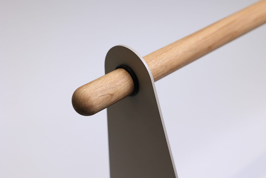 Nahaufnahme eines WoodUpp-Produkts: eine runde Eichenholzstange, sichtlich geölt, die durch eine matte, sandfarbene Metallplatte als Standfuß geführt wird. WoodUpp Garderobenstange kombiniert natürliche Holzmaserung und minimalistisches Metallträger-Design, mit Gummiring als stoßdämpfende Verbindung für Handtücher oder Kleidung geeignet, präzise verarbeitet und ästhetisch schlicht.