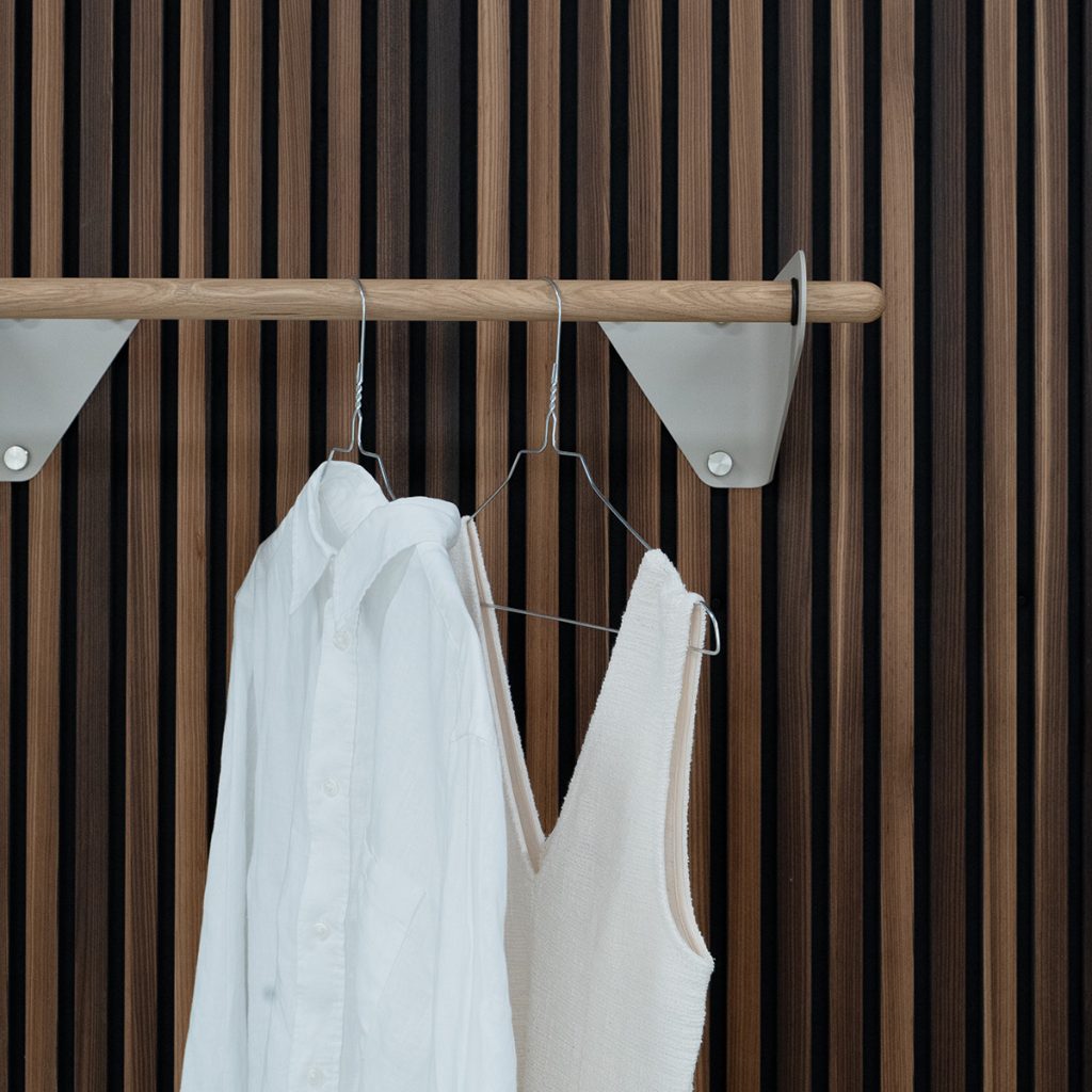 WoodUpp zeigt eine dunkel-helle Lamellenholz-Wand aus kontrastierendem Nuss- und Eichenfurnier mit geölter Eichenstange als Kleiderstange, gehalten von sandfarbenen, dreieckigen Metall-Wandhaltern. An der Stange hängen Drahtbügel mit einer weißen Hemdbluse und einem cremefarbenen Stricktop, im skandinavischen, warmen Design, ideal für Flur oder Schlafzimmer.