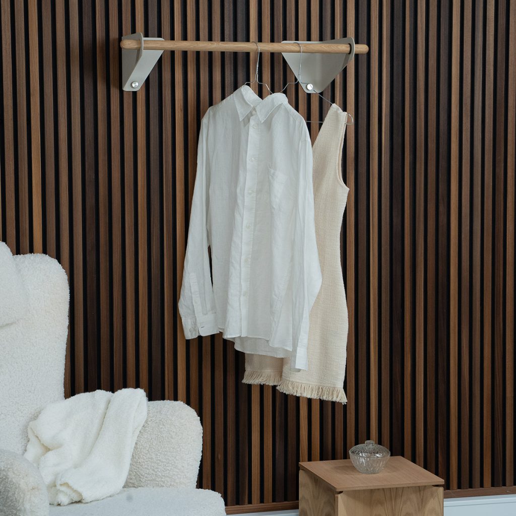 WoodUpp Wandgarderobe mit runder Eichenholzstange und zwei sandfarbenen, dreieckigen Metallhalterungen, an der eine weiße Leinenbluse und ein beigefarbenes, grobgewebtes Kleid mit Fransen hängen. Ergänzt durch einen kleinen WoodUpp Beistelltisch aus hellem Holz mit Glasschale.