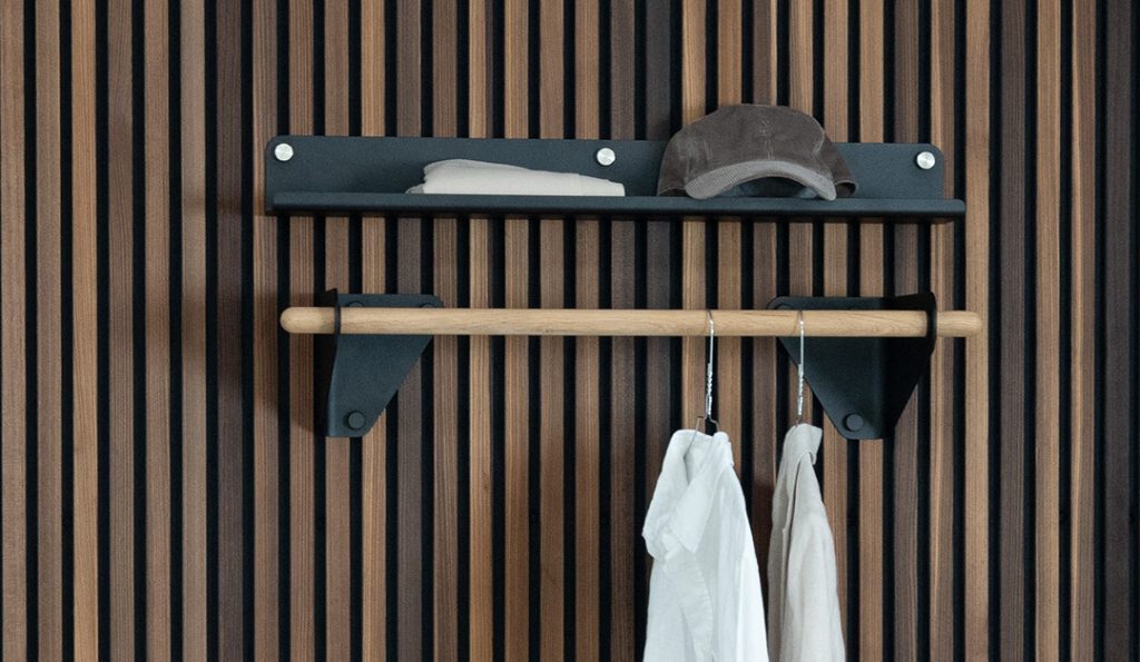 WoodUpp Wandgarderobe: skandinavisch inspirierte, minimalistische, schlanke Eichenstange als Kleiderstange, geölt und gehalten von mattschwarzen Metallwinkeln mit sichtbaren Schrauben. Darüber ein flacher schwarzer Ablageboden mit Kappe und gefaltetem Tuch. Zwei weiße Hemden an Metallbügeln hängen vor einer elegant lamellierten, vertikalen Holzpaneelwand.