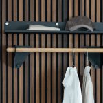 WoodUpp Wandgarderobe: skandinavisch inspirierte, minimalistische, schlanke Eichenstange als Kleiderstange, geölt und gehalten von mattschwarzen Metallwinkeln mit sichtbaren Schrauben. Darüber ein flacher schwarzer Ablageboden mit Kappe und gefaltetem Tuch. Zwei weiße Hemden an Metallbügeln hängen vor einer elegant lamellierten, vertikalen Holzpaneelwand.