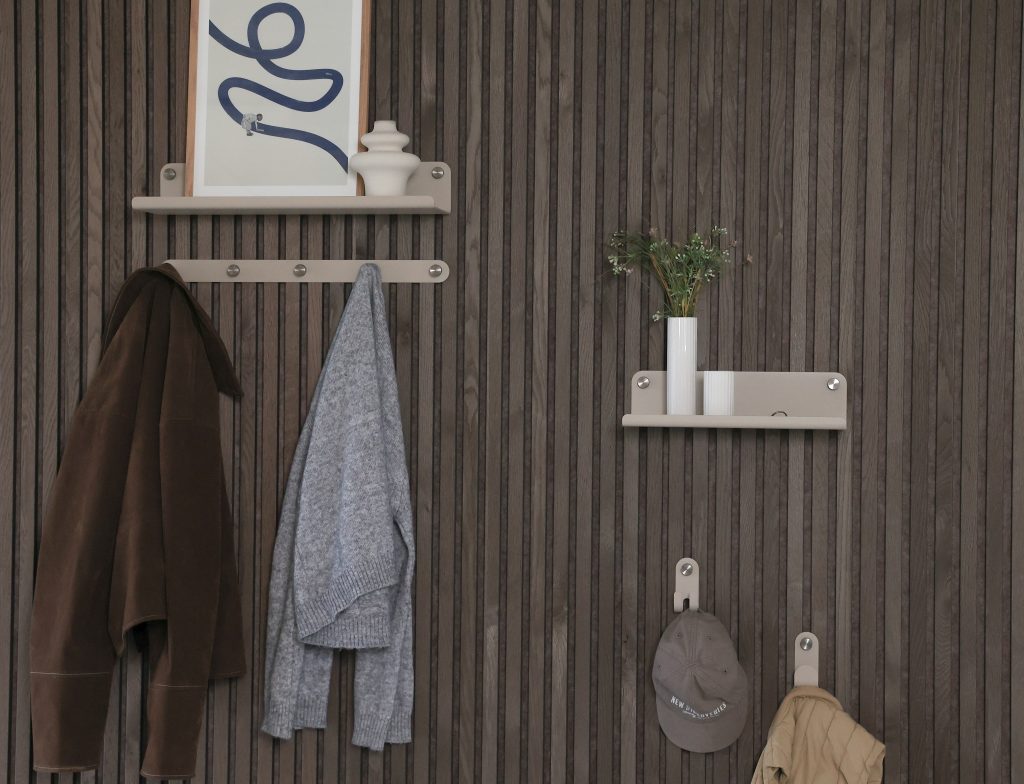 An dunkler Lamellenwand sind WoodUpp Wandmodule in Beige (Regal, Garderobenleiste und Einzelnhaken) montiert: oben ein kleines Regal mit Bild und Vase, links eine Hakenleiste mit drei Metallknöpfen trägt Jacke und Pullover, rechts ein Regal mit Vase sowie zwei einzelne Haken mit Cap und Jacke.