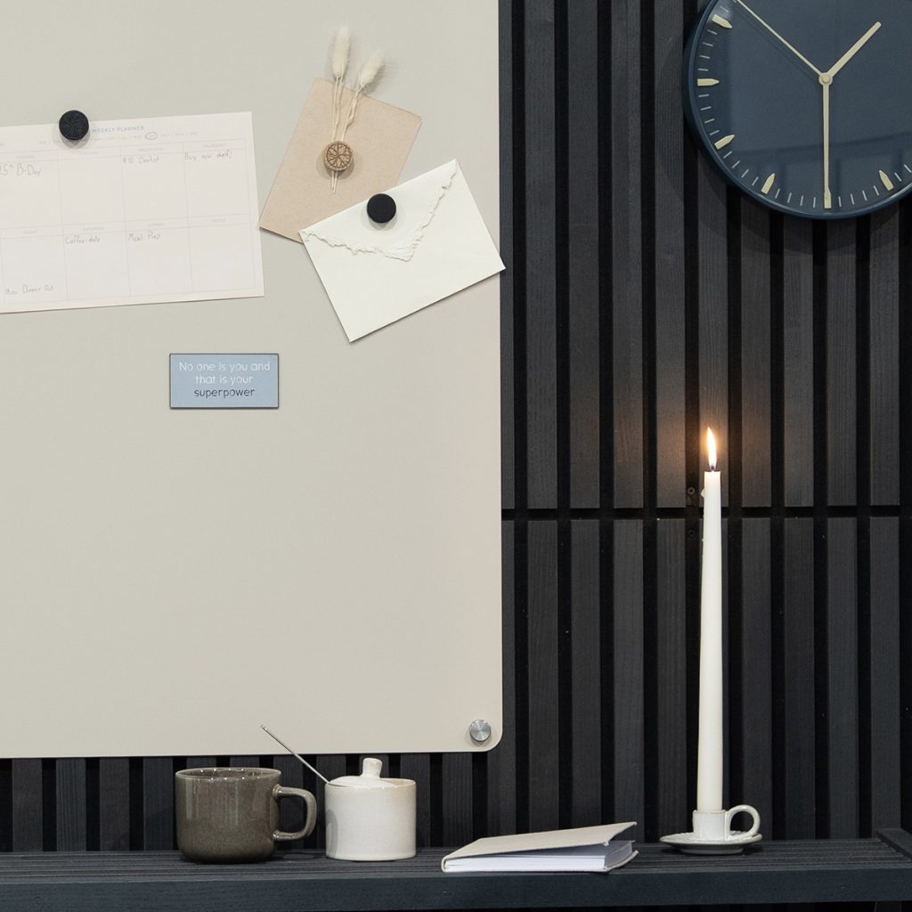 WoodUpp-Pinnwand: helle magnetische Tafel mit runden Magneten, befestigtem Wochenplanerblatt, Briefumschlag, getrockneten Deko-Gräsern und kleinem blauen WoodUpp-Quote-Magnet. Rechts dunkle WoodUpp-Lamellenholzverkleidung mit schlichter Wanduhr. Auf Regal brennende Kerze, Tasse, Zuckerdose und geschlossenes Notizbuch, ruhige Farbpalette.