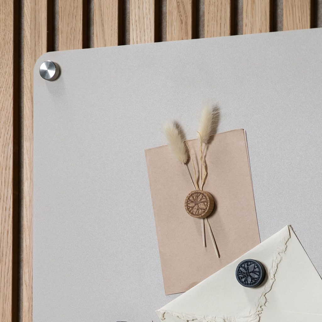WoodUpp Magnettafel in hellem Grau vor vertikalen Eichenlamellen, befestigt mit silbernem Metallabstandshalter; darauf ein helles, rundes WoodUpp-Holzmagnet mit filigraner Gravur und getrockneten Lagurus-Blüten an einer Kraftpapier-Karte, außerdem ein dunkler WoodUpp-Magnet als Briefsiegel auf einem rauen Umschlag im minimalistischen Stil mit natürlichen Farbtönen.