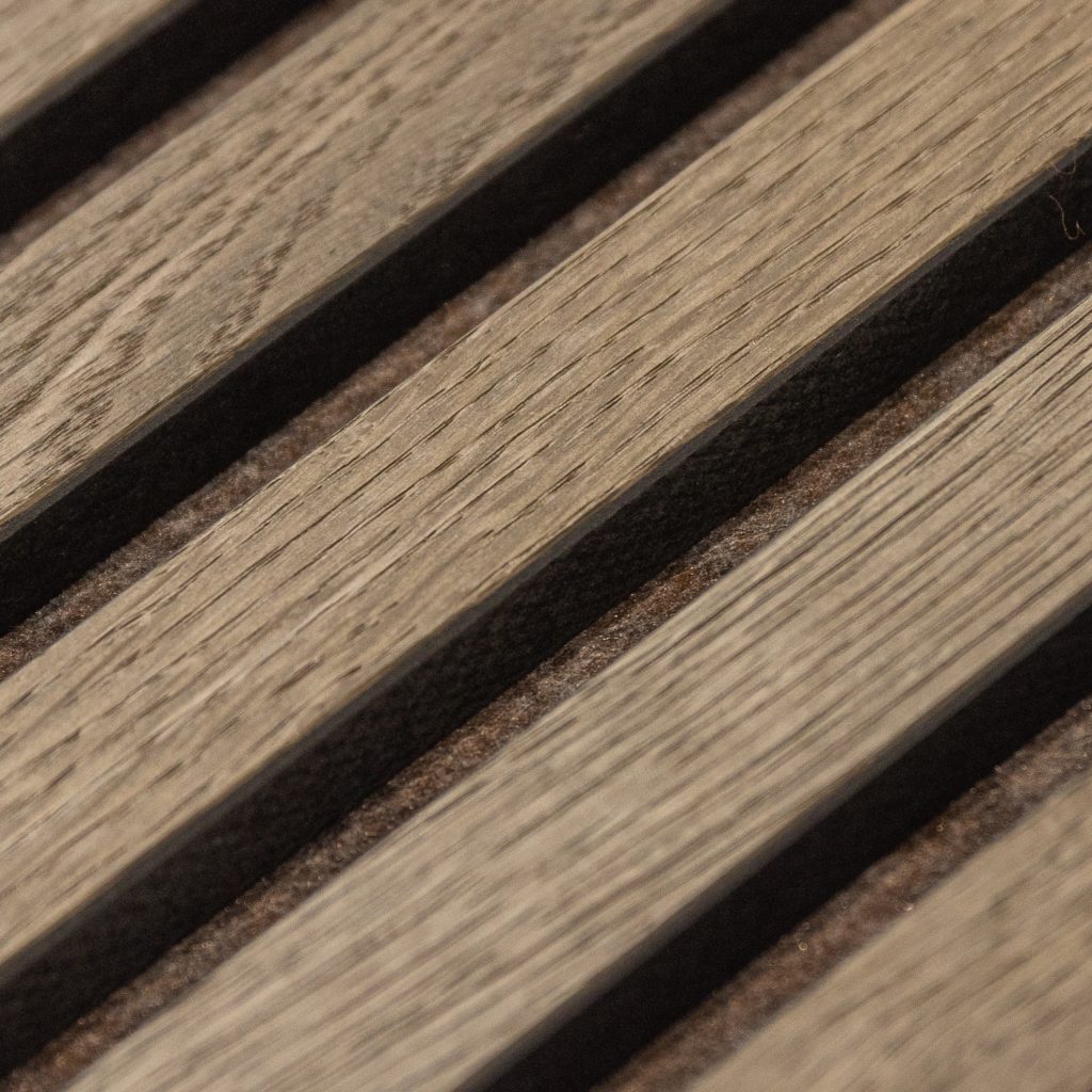 Nahe Aufnahme eines WoodUpp Akustikpaneels: diagonal angeordnete, schmale Holzlamellen mit sichtbarer Holzmaserung und matter, leicht gebürsteter Oberfläche in warmgrau-braunen Tönen, zwischen denen matte, dunkelbraune Filzstreifen als Akustikzwischenlagen sitzen. Man erkennt feine Jahresringe und die natürliche Textur. Die Lamellen bilden schmale Fugen und zeigen Detailstruktur der WoodUpp-Produkte.