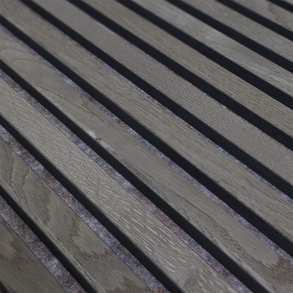 Nahaufnahme von WoodUpp Akustiklamellen: gleichmäßig angeordnete, helle Eichenholzlatten in diagonaler Perspektive mit sichtbarer Maserung und schmalen schwarzen Fugen, hinterlegt mit graubraunem Akustikvlies. Das Bild zeigt die strukturierte Oberfläche und den funktionalen Abstand der WoodUpp Lamellenpaneele für Raumakustik und Design, ästhetisch und funktional.