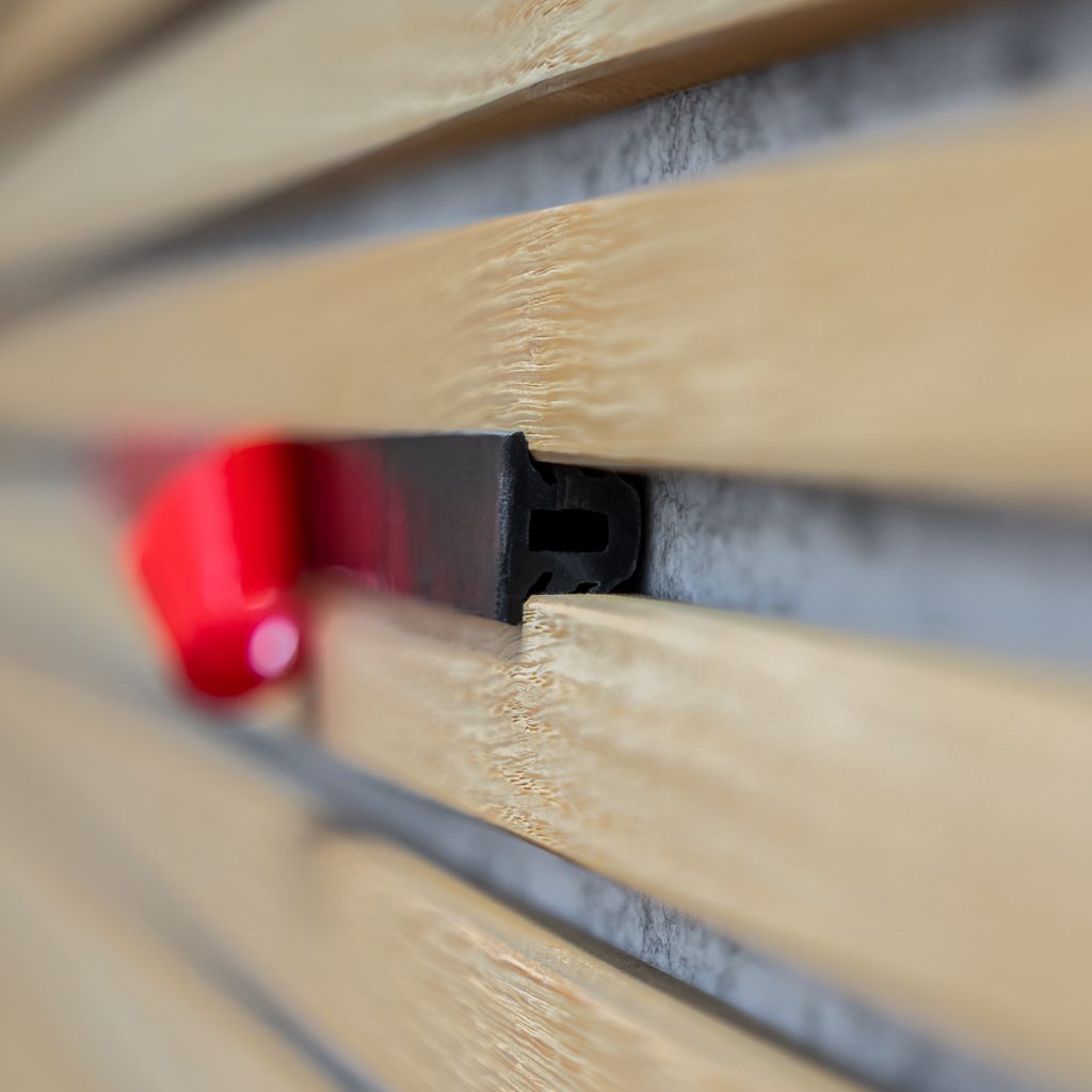 Nahaufnahme einer WoodUpp-Wandverkleidung: helle, naturfarbene Holzlamellen mit deutlicher Maserung, eng angeordnet. Dazwischen sitzt ein schwarzes WoodUpp-Montageprofil/Montageclip mit rechteckigem Hohlraum; im unscharfen Hintergrund ein roter Verriegelungsclip. Geringe Tiefenschärfe betont das profilierte Kunststoffteil und die präzise Passform und zeigt das einfache Montageprinzip der WoodUpp-Elemente an der Wand.