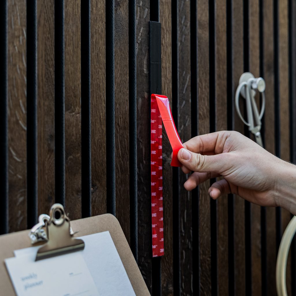 Eine Hand zieht die glänzende rote 3M-Trägerfolie eines WoodUpp Montageklebebands ab, um eine schlanke, schwarze WoodUpp Magnetleiste mit rechteckigem Profil an einer dunklen Holzlamellenwand zu befestigen. Im Bild sind außerdem Klemmbrett, Metallhaken und Kabelhalter zu sehen, Fokus auf Leiste und Klebestreifen.