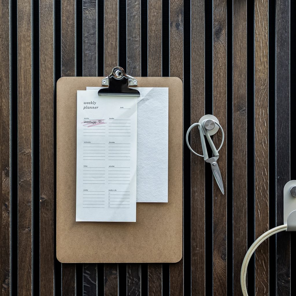WoodUpp: Senkrechte, schmale dunkelbraune Holzlamellenwand mit kontrastreichen schwarzen Fugen und sichtbarer Maserung als Hintergrund. Davor ein WoodUpp-Klemmbrett aus MDF mit chromierter Metallklammer und befestigtem weißem Wochenplaner-Block mit feinem Linienlayout. Rechts hängt eine silberne Schere an rundem Magnethaken, dazu praktischer Kabelhalter.