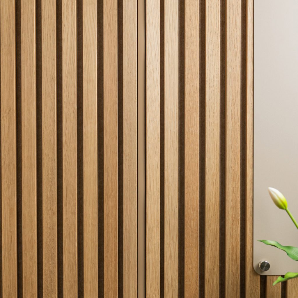 WoodUpp lamelpaneler i lyst, naturligt egetræ dækker billedets flade med jævne, lodrette lister og mørke mellemrum; til højre ses en elegant WoodUpp beige opslagstavle/plade med diskret synlig rund metalskrue og i nederste højre hjørne en enkelt tulipanknop og grønne blade.