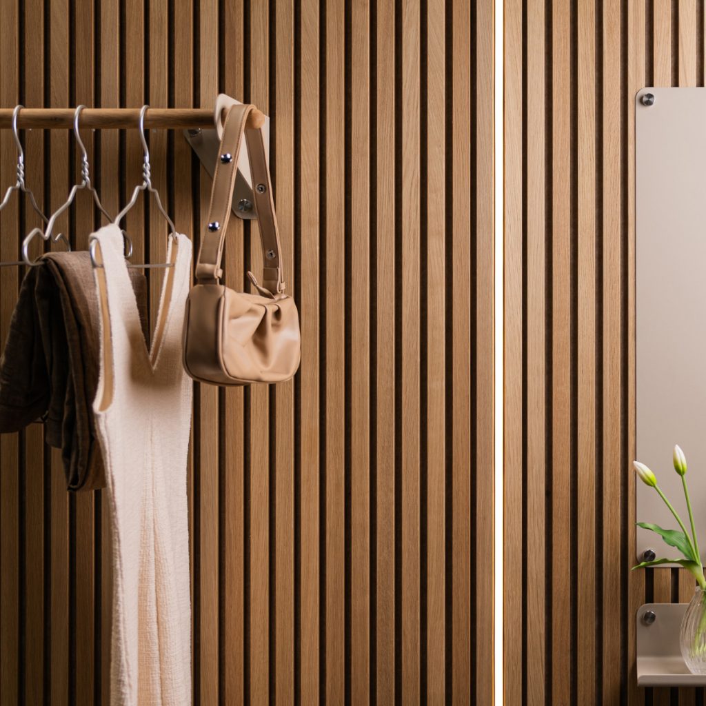 WoodUpp lamelvæg i varm, naturlig træfinish danner baggrund for en WoodUpp træ-tøjstang monteret med metalbeslag, metalbøjler og hængende tøj samt lyse tasker. Til højre ses et WoodUpp vægspejl med små hylder og en glasvase, alt i enkelt, moderne, stilrent design.