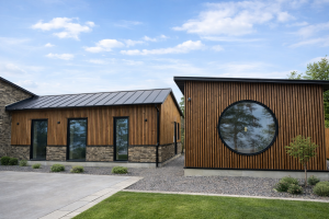 Udvendigt billede af moderne bygninger med facadebeklædning fra WoodUpp. De lodrette trælameller i en varm, gyldenbrun farve dækker den ene bygning helt omkring et stort rundt vindue, og den øvre del af den anden bygning over en stensokkel.