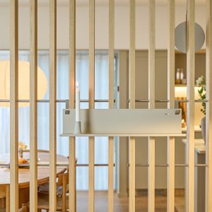 WoodUpp: Schmale, sandfarbene Metallablage mit integriertem Kerzenhalter, an einem hellen Eichenlamellen-Raumteiler montiert. Die flache, matte Ablage zeigt minimalistisches Design; darauf steht eine brennende weiße Stabkerze. Im Hintergrund entspannter Essbereich mit rundem Holztisch, Papierleuchte und sanfter natürlicher Beleuchtung als dezenter Dekoakzent für ein luftiges Raumgefühl.