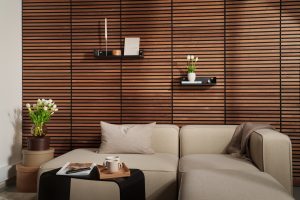 Modernes Wohnzimmer mit WoodUpp-Holzlamellenwand in warmem Walnusston mit schwarzen Horizontalfugen; zwei schmale, matte WoodUpp-Wandregale mit runden Messingbefestigungen, bestückt mit Kerzenständer, Buch, Holzschale, Vase mit weißen Tulpen sowie kleiner Deko; hellbeige Eckcouch, Beistelltisch, Magazin, Tasse und Plaid vervollständigen die wohnliche Szene.