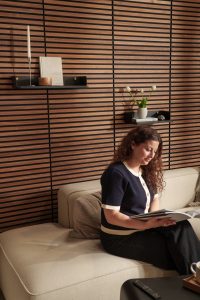 WoodUpp-Wandverkleidung aus horizontalen Holzlamellen bildet den Hintergrund; zwei schwarze, schmale WoodUpp-Schweberegale mit goldfarbenen Befestigungen tragen Kerze, Buch, Vase mit drei Tulpen und eine kleine Dekorkugel, elegant und minimalistisch gestaltet. Vor der Wand sitzt eine Frau auf hellem Sofa und liest ein Buch.