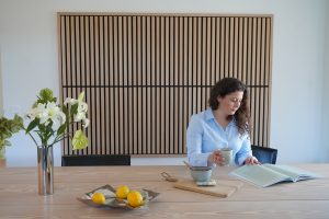 Eine Frau sitzt an einem eleganten, hellen WoodUpp Holztisch vor einem großen WoodUpp Lamellen-Akustikpaneel aus Eichenleisten mit schwarzem Hintergrund. Auf dem Tisch liegen Keramikschale, Schneidebrett, Zitronen in einer Schale und eine Vase mit Blumen; sie hält eine Tasse und liest ein Buch.