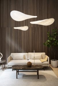 Modernes Wohnzimmer mit WoodUpp-Lamellenwand aus dunklen vertikalen Holzpaneelen im Hintergrund. Davor beige Dreisitzer-Couch mit Kissen und senfgelbem Plaid, dunkel marmorierter Couchtisch mit runder Vase und Kerzenleuchter, zart leuchtende, tropfenförmige Hängeleuchten, Topfpflanze rechts, heller Teppich und kleine Skulptur links. WoodUpp-Paneele setzen warmen, strukturierten Akzent.