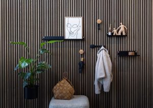 Eleganter Eingangsbereich mit WoodUpp-Wandpaneelen und modularen WoodUpp-Produkten: mattschwarze, schmale Wandregale mit sichtbaren Messingschrauben, runde, eichenfarbene Holz-Kleiderhaken und mattschwarze Hakenhalter. Auf Ablagen gerahmtes Linienbild, Dekofiguren, Duftstäbchen; an Haken Mantel, Regenschirm, geflochtener Korb und große Topfpflanze. Sitzbank mit beigem, strukturiertem Polster unterstreicht warmen, skandinavischen Stil.