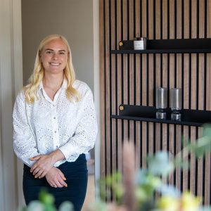 Porträt einer lächelnden Frau neben einer WoodUpp-Lamellenwand aus warmem Holz mit zwei schwarzen WoodUpp-Wandregalen, goldenen Befestigungen und darauf zwei gerippten Gläsern sowie einer Duftkerze; Vordergrund mit unscharfen Pflanzen. Lamellen vertikal, Regale matt schwarz mit runden Messingknöpfen, Frau in weißer Bluse.