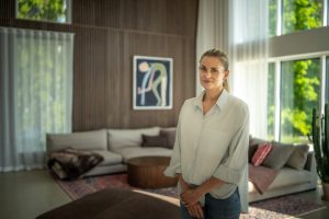 Modernes Wohnzimmer mit einer Frau vor einer WoodUpp-Holzlamellenwand aus feinen, senkrechten Holzlamellen und einem runden WoodUpp-Couchtisch mit geriffelter, vertikaler Rippenoptik. Beide WoodUpp-Produkte zeigen warme Eiche- oder Nussbaumtöne, natürliche Maserung und klaren, minimalistischen Designcharakter; dazu Sofa, Teppich und viel Tageslicht.