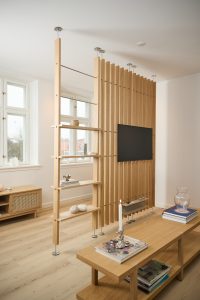 WoodUpp Raumteiler mit schlanken, vertikalen Eichenlamellen und integrierten Regalböden sowie TV‑Halterung, gespannt zwischen Boden und Decke mit Edelstahlbefestigungen. Dazu ein niedriger WoodUpp Couchtisch und Sideboard in hellem Eichenholz, schlichtes skandinavisches Design, offene Ablagen mit Büchern und Deko, natürlichem Ton und Kerzenlicht.