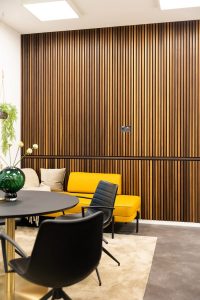 Moderner Loungebereich mit WoodUpp-Holzlamellenwand: warme, vertikale Holzlamellen in walnussfarbener Optik mit schmalen, dunklen Fugen, gleichmäßig profiliert und matt. Sichtbar sind Steckdosen in der Platte. Im Vordergrund gelbe Sitzbank, schwarze Stühle, runder Tisch, grüne Vase und Teppich. WoodUpp-Wand schafft akustische, dekorative Atmosphäre.