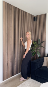 Eine lächelnde Frau präsentiert eine stilvolle Raumecke, die mit WoodUpp Akustikpaneelen in Walnuss verkleidet ist. Die dunklen Holzlamellen auf schwarzem Filz reichen vom Boden bis zur Decke und bilden eine moderne Holzwand hinter einer großen Pflanze und einem Sofa.