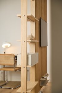 Nahaufnahme eines WoodUpp Raumteilers aus hellem Eichenholz mit schmalen Regalbrettern, einer montierten metallischen Aufbewahrungsbox und einer kleinen Dekorrolle. Rechts sichtbar: an der Rückseite befestigter TV-Bildschirm; im Hintergrund Schreibtisch, Stuhl und Lampe. Die sichtbaren Schrauben und Metallbeschläge betonen das modulare, minimalistische Design von WoodUpp.