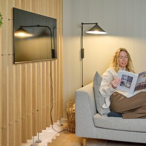 Gemütliches Wohnzimmer mit Sofa und entspannter Frau, die eine Zeitschrift liest. Links ist eine WoodUpp-Holzlamellen-Wandverkleidung in natürlichem Eichenholzton mit schmalen vertikalen Lamellen zu sehen. Die WoodUpp-Lamellenpaneele stehen auf sichtbaren Metallfüßen; außerdem sind Fernseher, Kabel, Wandlampe und Korb erkennbar.