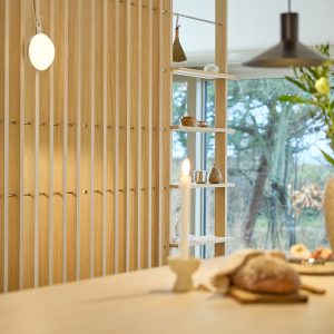 WoodUpp Holzlamellen-Trennwand aus hellen Eichenleisten mit eingelassenen Metallhaken und schmalen, höhenverstellbaren Einlegeböden, auf denen Keramikschalen, kleine Dekoobjekte und ein Mini-Besen stehen. Im Vordergrund brennende Kerze und frisches Brot auf dem Tisch, im Hintergrund Fenster mit Blick ins Grüne.