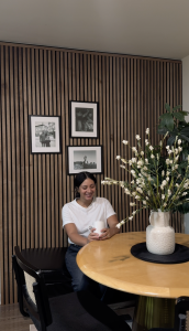 Eine lächelnde Frau sitzt an einem runden Holztisch und hält eine weiße Tasse. Hinter ihr ist eine Wand, die vollständig mit WoodUpp Akustikpaneelen in Eiche verkleidet ist. Die vertikalen Holzlamellen auf schwarzem Filz schaffen eine warme, stilvolle Kulisse und sind mit drei gerahmten Schwarz-Weiß-Bildern dekoriert.