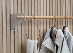 WoodUpp Wandpaneel mit vertikalen Eichenlamellen, darauf eine WoodUpp Garderobenstange aus massivem Eichenholz, gehalten von einem dezenten, sandgrauen Metall-Winkelträger. An der Stange hängen filigrane Lederbügel, ein Leder-Schalhalter und zwei helle Mäntel. Der runde Holzstab zeigt feine Maserung, Befestigungsschraube sichtbar, minimalistische Ästhetik.
